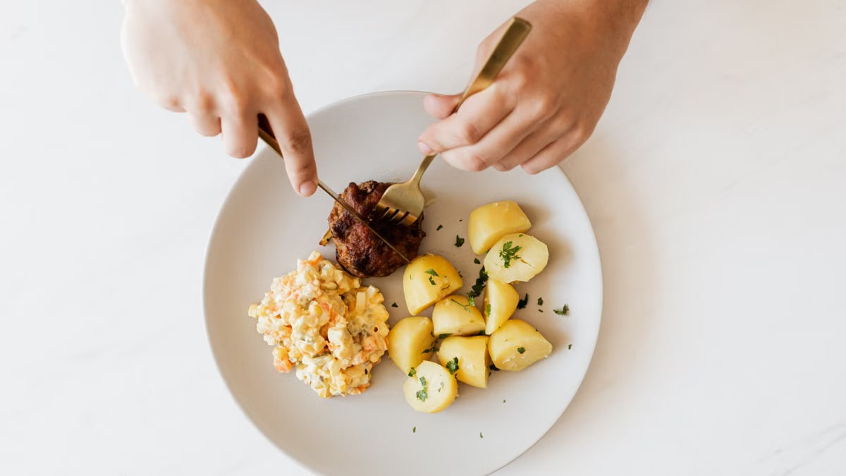 Top view of crop unrecognizable male with cutleries slicing delicious beef cutlet garnished with boiled potatoes and parsley and portion of Russian meat salad on plate placed on white table